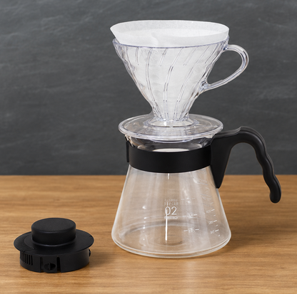 A clear glass pour-over coffee maker with a white paper filter cone, black handle, and black collar, shown on a wooden surface against a dark gray background. A separate black lid is placed to the left.