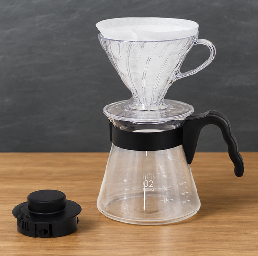 A clear glass pour-over coffee maker with a white paper filter cone, black handle, and black collar, shown on a wooden surface against a dark gray background. A separate black lid is placed to the left.